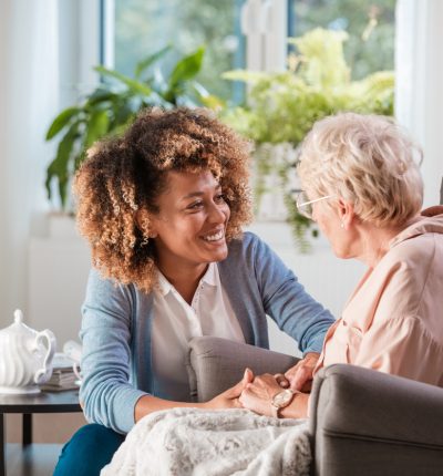 Female home caregiver talking with senior woman, sitting in living room and listening to her carefully.